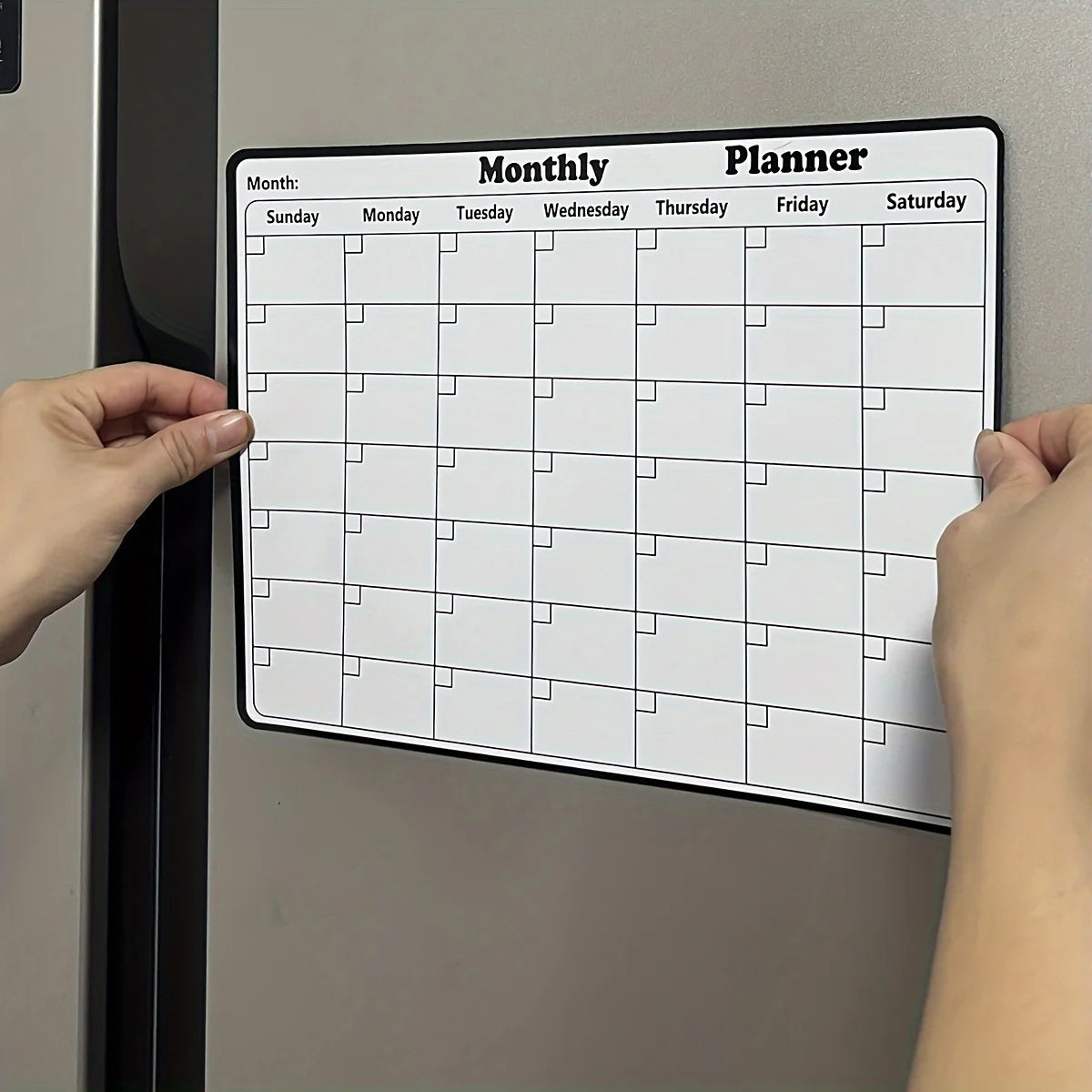 A hand holds a magnetic dry erase monthly calendar planner for the refrigerator with grid lines and days of the week.
