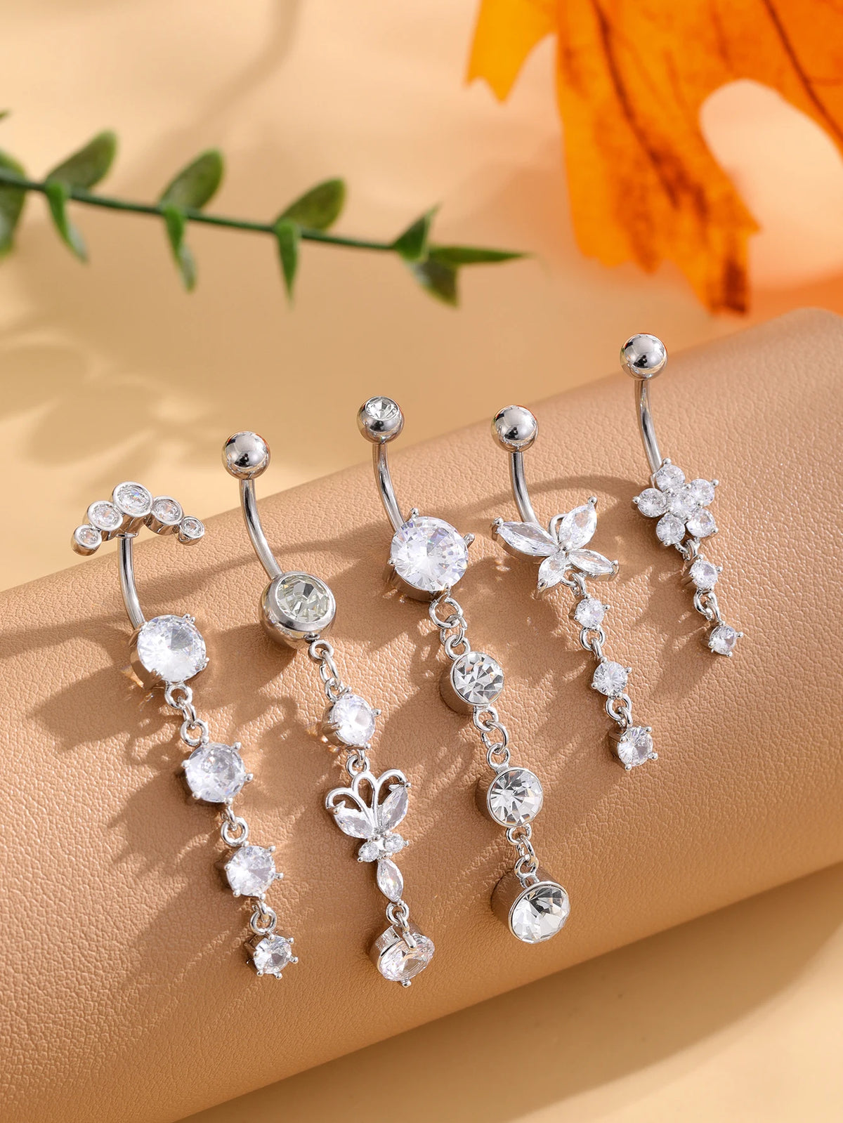 A set of five gold tone butterfly and flower belly rings with clear stones displayed on a beige surface.