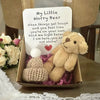 A small, light brown teddy bear plush with a handwritten card inside a cardboard box.