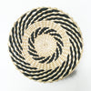 A round woven straw wall decor hanging with black and natural beige stripes in a spiral pattern.