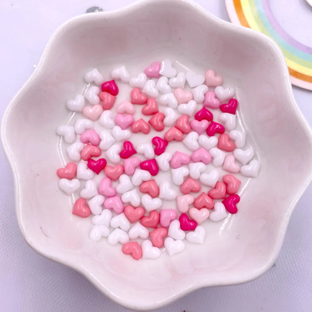 A small white dish filled with a mix of pink and white heart-shaped flatback rhinestones for nail art and crafts.