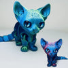 A blue and teal articulated 3D printed figurine of a Siamese cat with large eyes.
