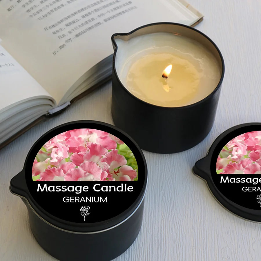 A black jar of Geranium massage candle with a lit wick sits next to an open book.