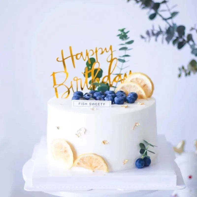 A white birthday cake decorated with gold lettering and fresh citrus slices, set against a light background.