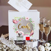 A white acrylic gift box with a floral wreath design and gold lettering, featuring a perfume bottle and a glass of amber liqu