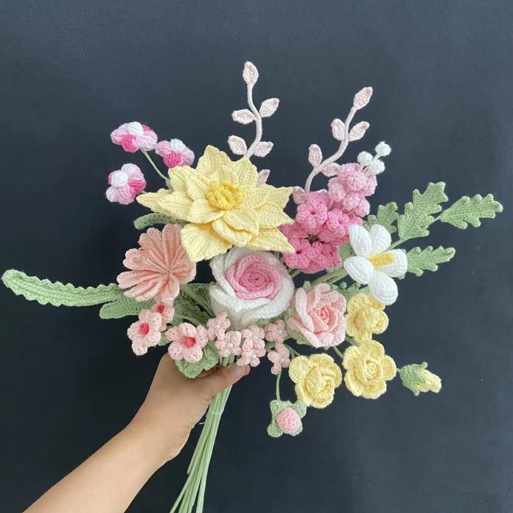 A hand holds a bouquet of handmade crochet flowers in shades of pink, yellow, and white.