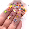 A hand displays various colorful 3D resin flower gems in shades of pink, purple, yellow, and white.