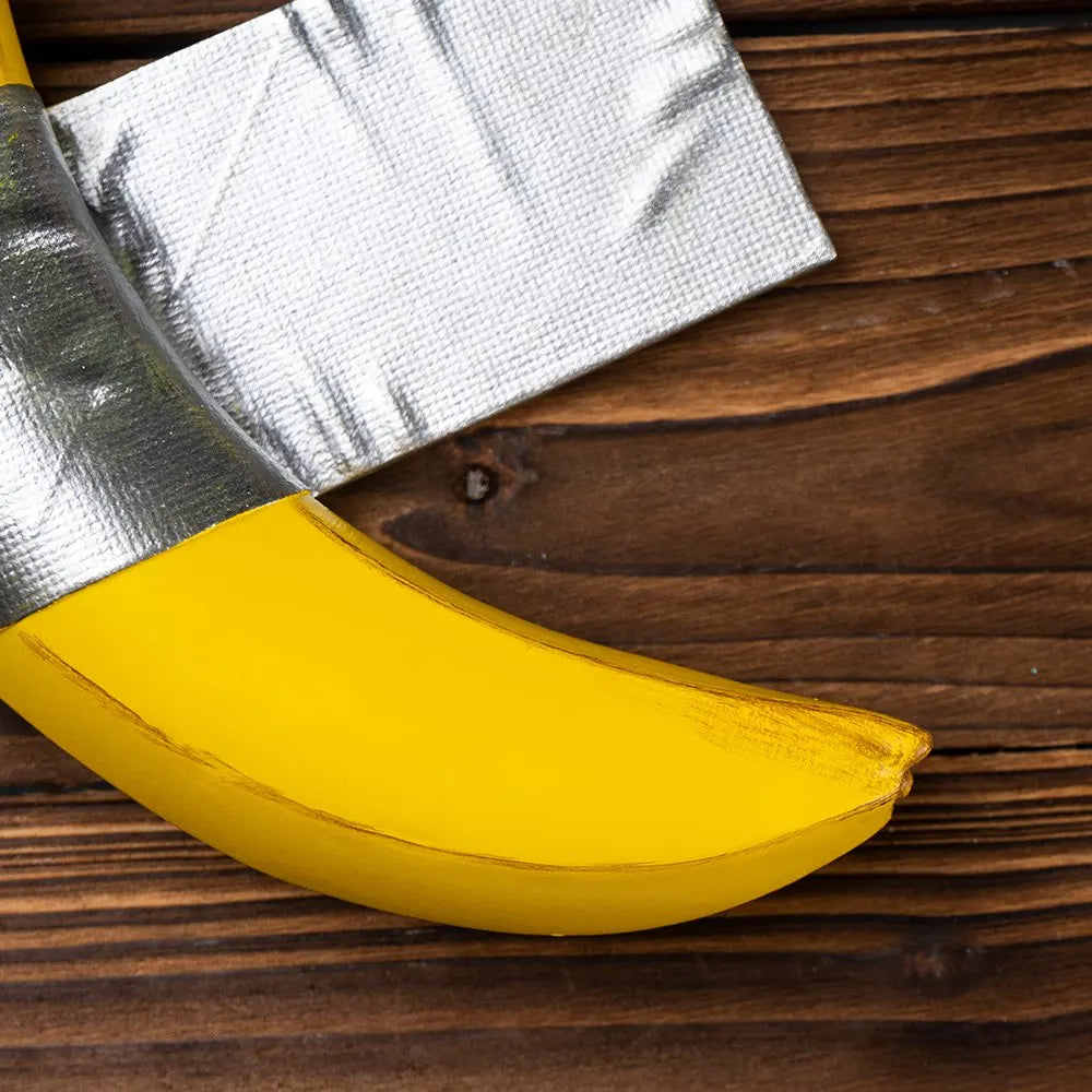 A yellow resin banana wall sculpture with a silver foil wrapper is shown on a dark wooden surface.