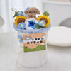 A handmade crochet flower bouquet featuring a brown bear and blue and yellow flowers displayed in a white vase.
