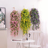 Three hanging silk morning glory vine flowers in pink, yellow-green, and purple on a white wall.