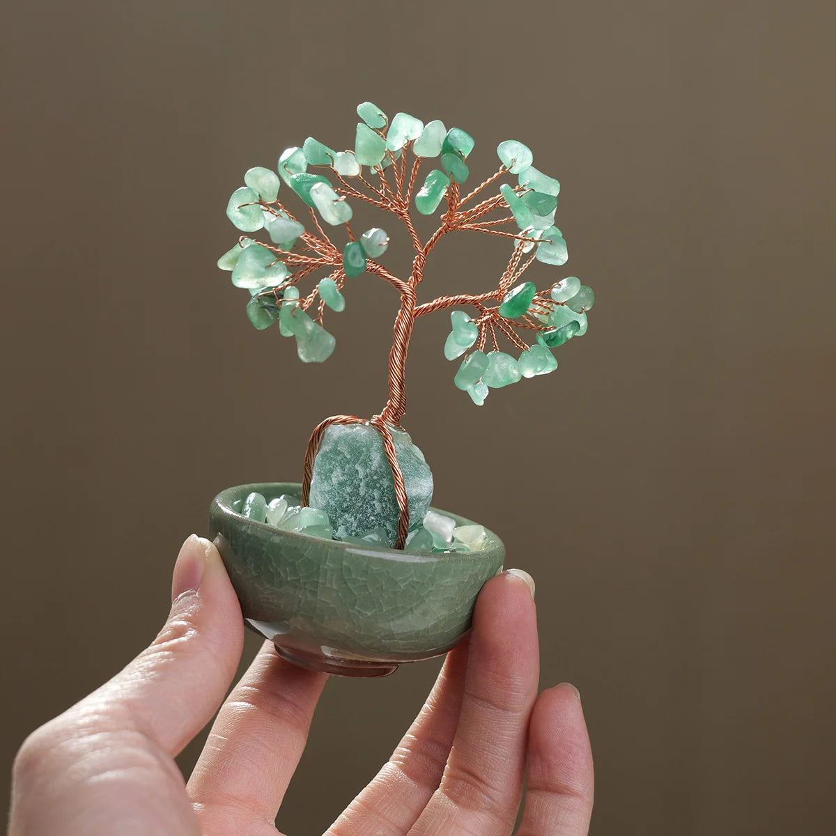 A hand holds a natural crystal money tree with rose quartz base featuring green crystals on a copper branch.