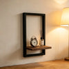 A black wooden floating wall shelf displaying a vintage alarm clock and a glass bottle with reeds.