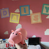 A hand holds an adorable pink piggy bank against a pink wall decorated with cloud shapes.