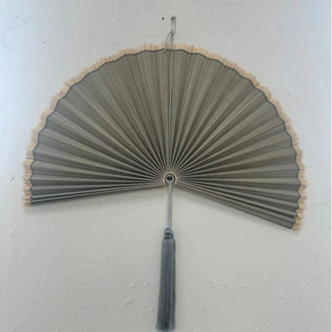 Handwoven bamboo wall fan decor with a light gray fan and tassel mounted on a white wall.