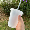 A hand holds a clear plastic cold cup with a lid and straw outdoors.