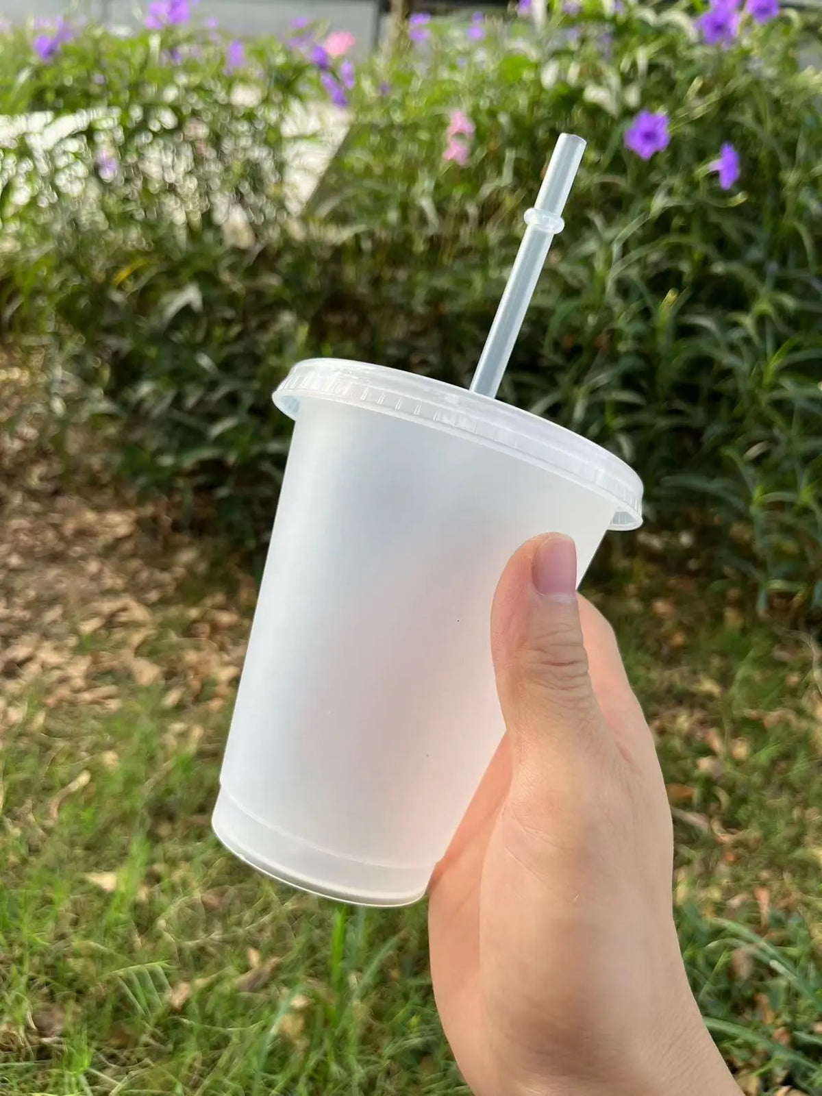 A hand holds a clear plastic cold cup with a lid and straw outdoors.