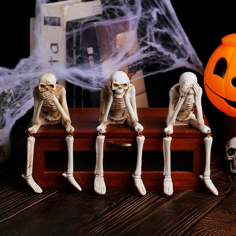 Three resin skeleton skull figurines seated on a wooden box with a white cobweb in the background.