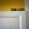 Un letrero de metal negro con la palabra THE LOO montado en una pared amarilla sobre un molde arquitectónico blanco.