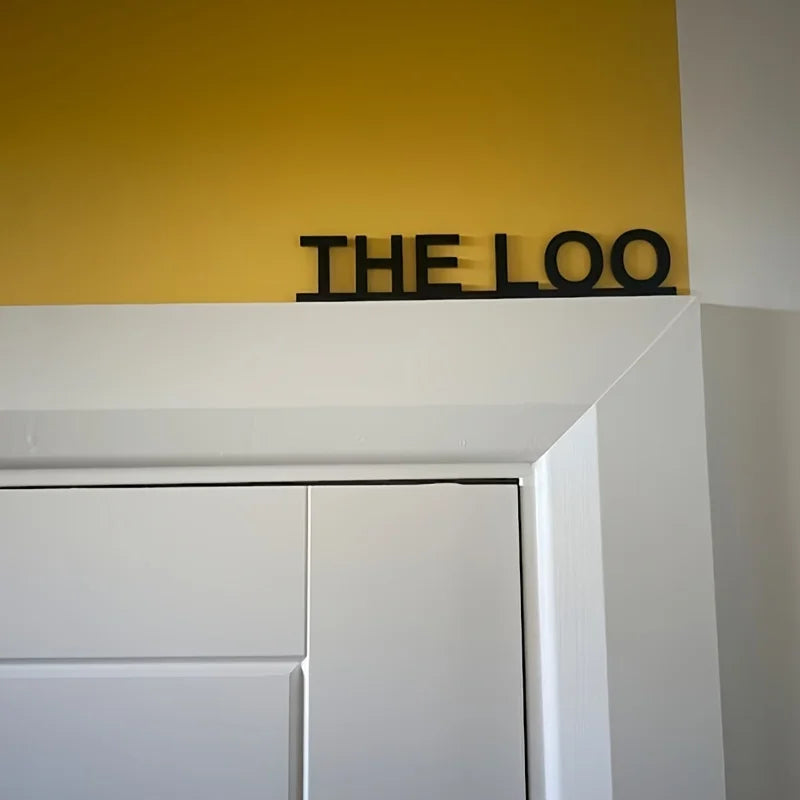 Un letrero de metal negro con la palabra THE LOO montado en una pared amarilla sobre un molde arquitectónico blanco.