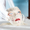 A pale, porcelain masquerade mask with delicate gray floral designs and red blood-like streaks on the face.