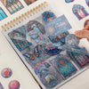 A hand is placing vintage floral window scrapbooking stickers into a spiral-bound notebook.