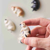 A hand holds a small cream-colored cat fridge magnet design against a light background with other cat designs visible.