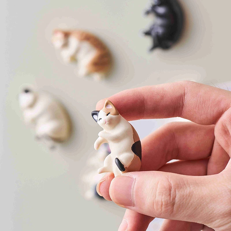 A hand holds a small cream-colored cat fridge magnet design against a light background with other cat designs visible.