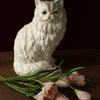 A white ceramic cat figurine sits next to a bunch of pink and white tulips on a brown surface.