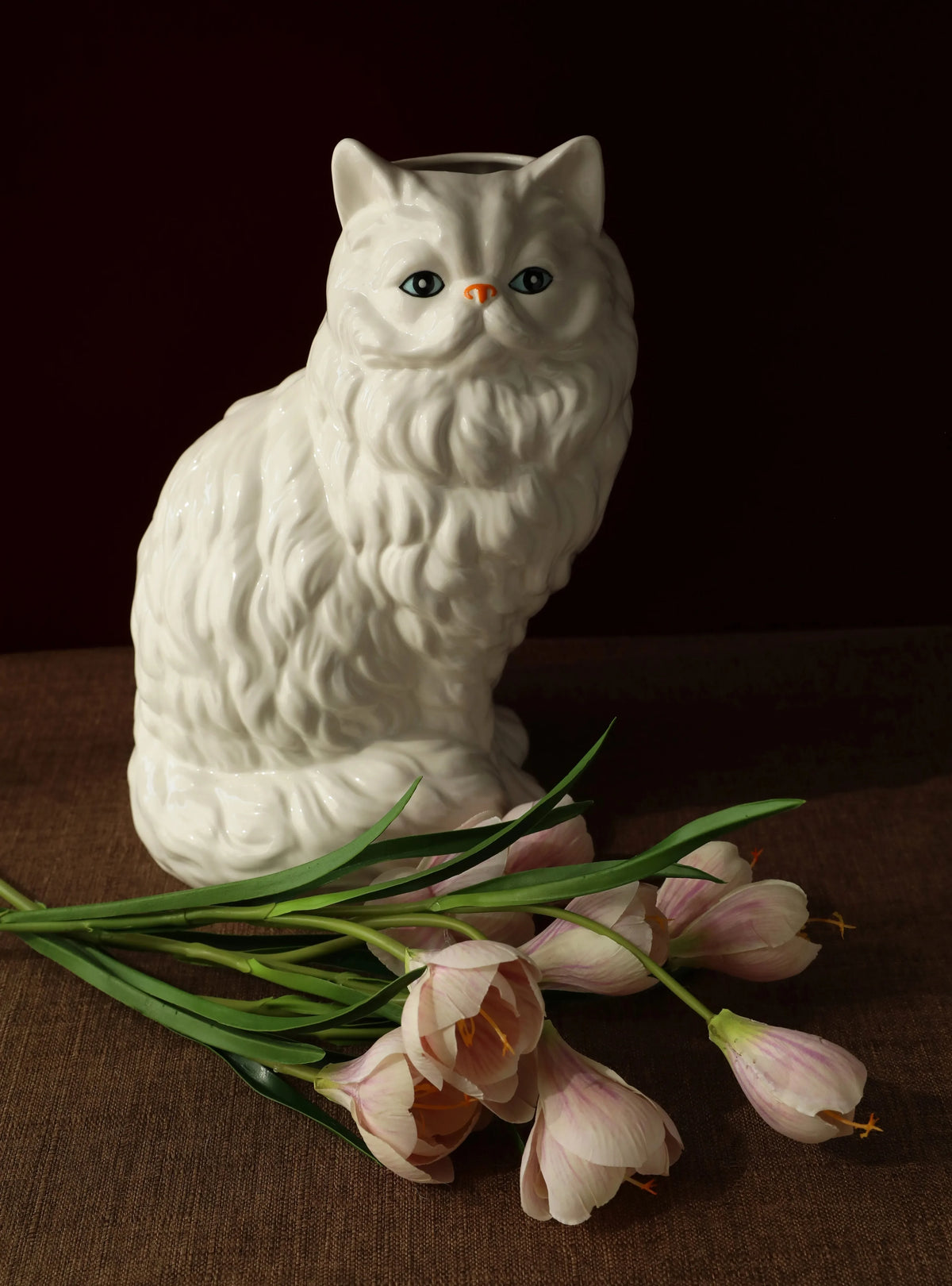 A white ceramic cat figurine sits next to a bunch of pink and white tulips on a brown surface.