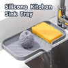 A silicone kitchen sink tray holds a yellow sponge and a scrubbing brush inside a stainless steel sink.