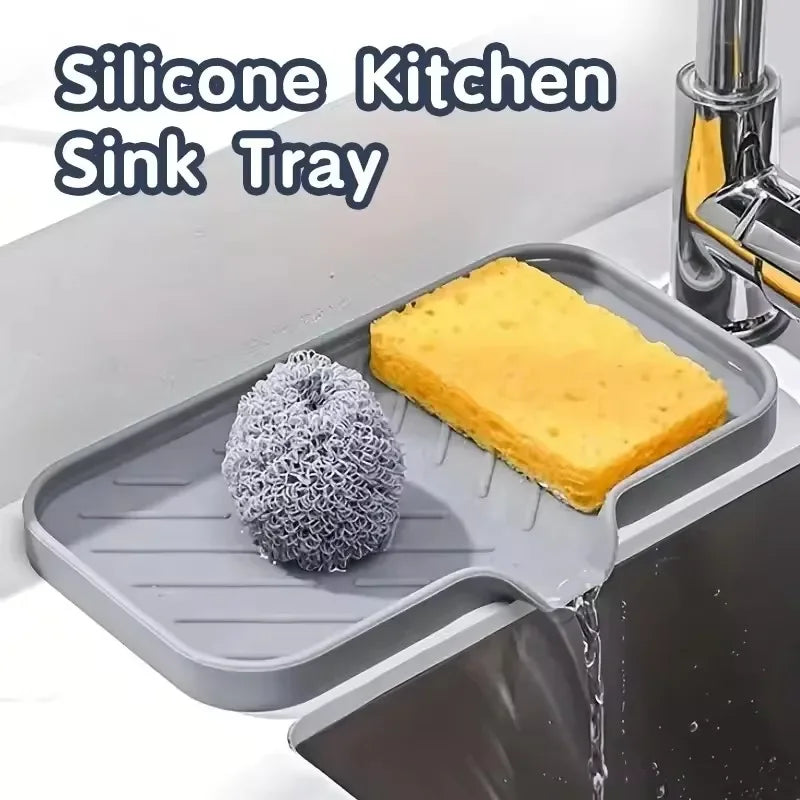 A silicone kitchen sink tray holds a yellow sponge and a scrubbing brush inside a stainless steel sink.