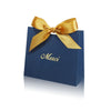 A dark blue paperboard gift bag tied with a golden ribbon, featuring the word Merci printed on the front.
