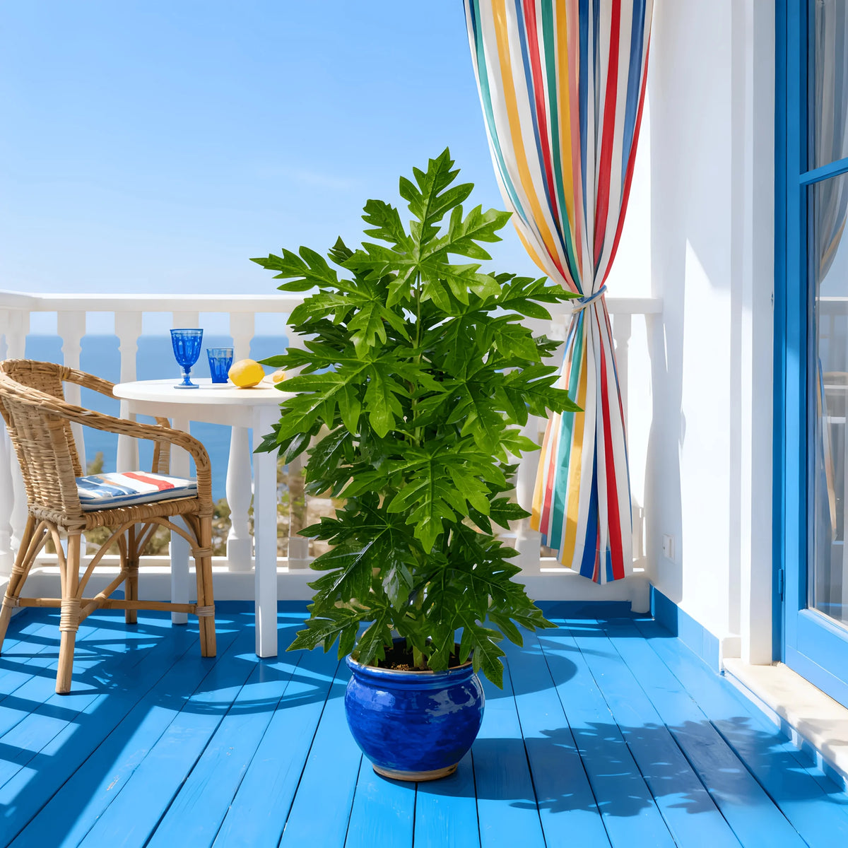 A vibrant green artificial papaya leaf plant in a blue pot sits on a wooden deck with a white railing and colorful curtains.