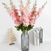 Un bouquet de fleurs artificielles de delphinium roses et blanches dans un vase en verre à côté d'une pile de livres.