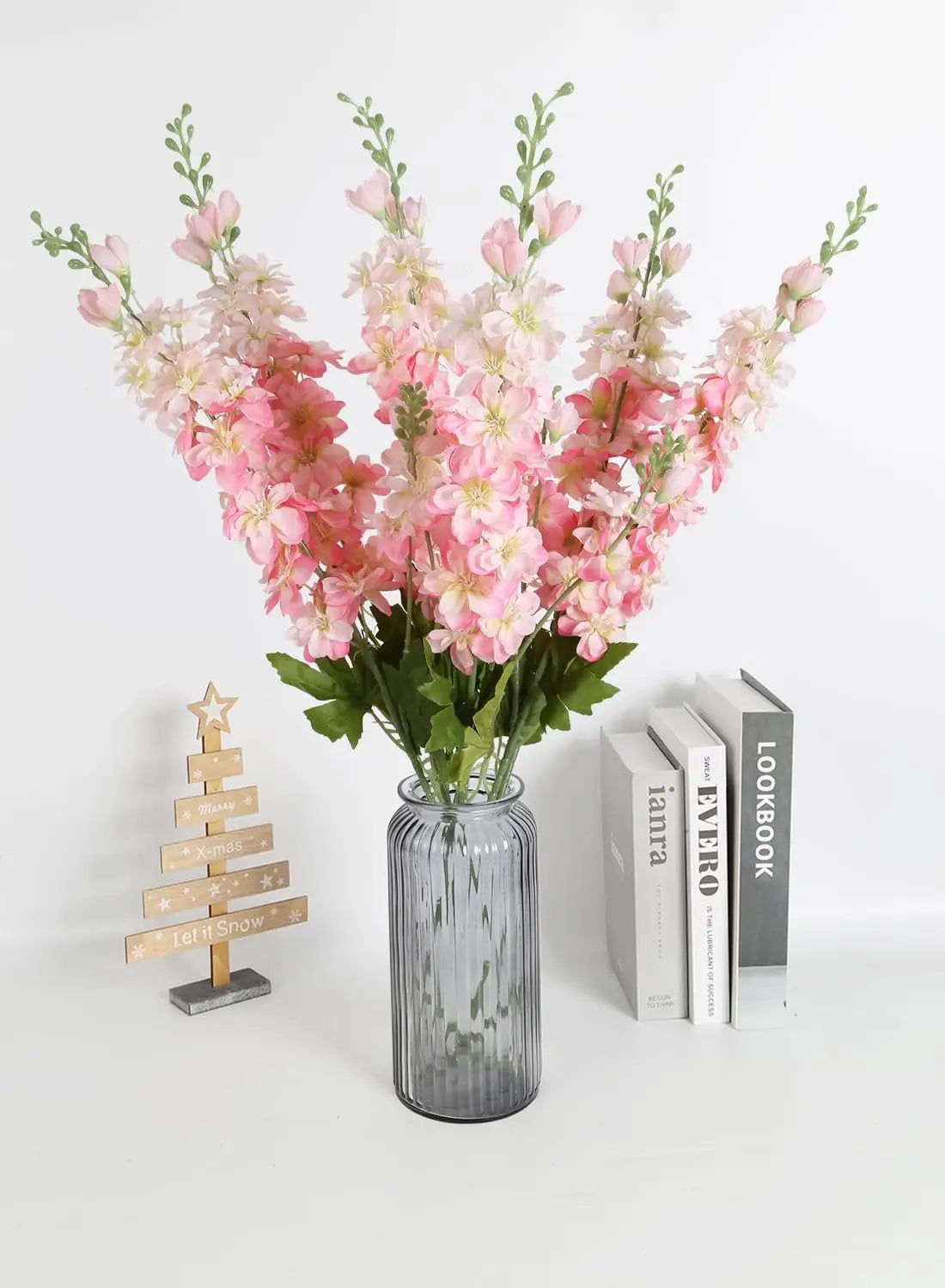 Un bouquet de fleurs artificielles de delphinium roses et blanches dans un vase en verre à côté d'une pile de livres.