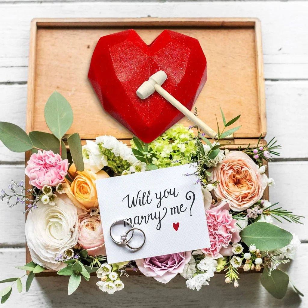 A red heart-shaped silicone mold with a hammer rests on a wooden tray surrounded by floral greenery.