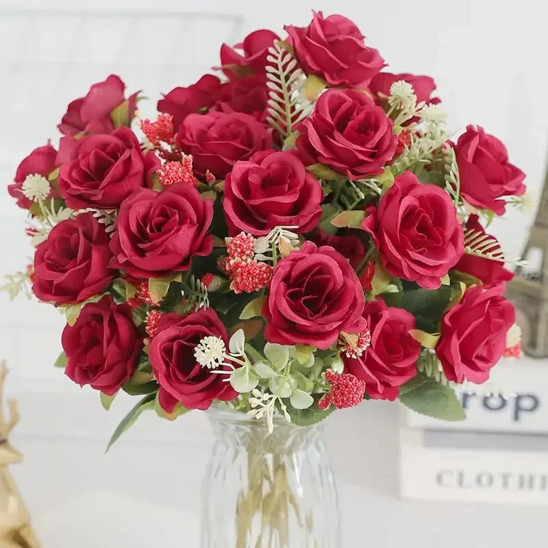 A bouquet of deep red silk artificial roses with green foliage and small pink accents in a clear glass vase.