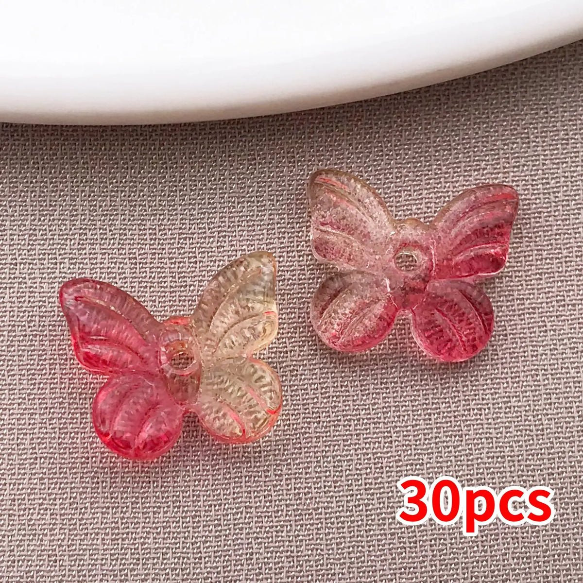 Un juego de 30 cuentas de mariposa de vidrio en tonos rosa y transparente, exhibidas sobre una superficie gris texturizada.