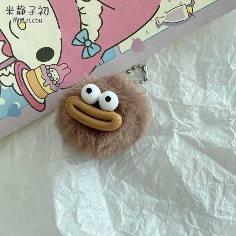 A small, fluffy brown plush charm shaped like a sausage with a wide, smiling mouth and large white eyes.