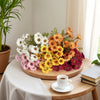 A wooden platter holds a vibrant arrangement of silk gerbera daisy artificial flowers in shades of white, yellow, orange, and