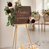 A wooden sign welcomes guests to a beginning decorated with artificial roses and greenery, set in a bright room.
