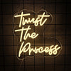 A glowing white neon sign spelling out Trust The Process is mounted on a dark grid wall.