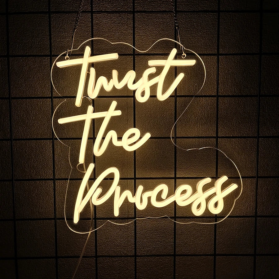 A glowing white neon sign spelling out Trust The Process is mounted on a dark grid wall.