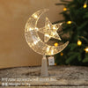 A glowing crescent moon and star ornament made of clear glass or acrylic, displayed in front of a green Christmas tree.