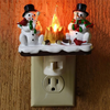 A decoration featuring two white snowmen with black hats and carrots, illuminated by a flickering orange campfire light.
