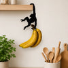 A black monkey-shaped storage hook is hanging on a shelf, positioned above a bunch of yellow bananas.