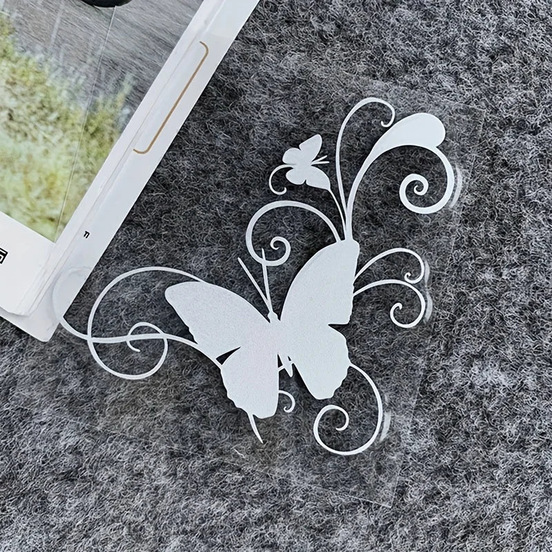 A white vinyl sticker of a butterfly with swirling vines and a small flower on a gray, textured surface.