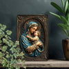 A bronze-colored metal sign depicting the Virgin Mary holding the infant Jesus in a detailed religious artwork.