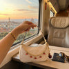 A hand holds a beige leather jewelry bag with a gold chain and red charms against a sunset view from a train window.