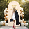 A woman in a white dress stands in front of a gold arch backdrop decorated with balloons and a graduation cap silhouette.